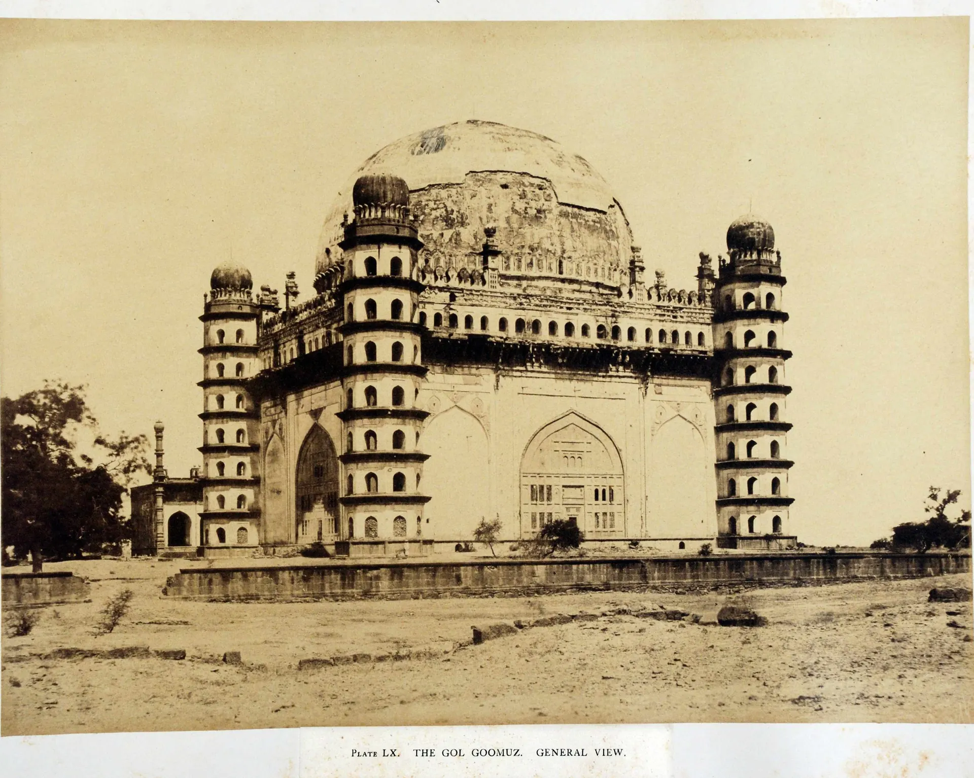 Sepia photograph of a massive cubical tomb with a great hemispherical dome and seven-storeyed octagonal towers at the corners, standing on a bare plain.