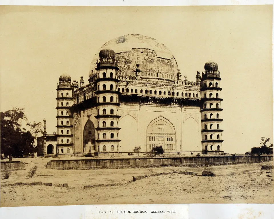Sepia photograph of a massive cubical tomb with a great hemispherical dome and seven-storeyed octagonal towers at the corners, standing on a bare plain.