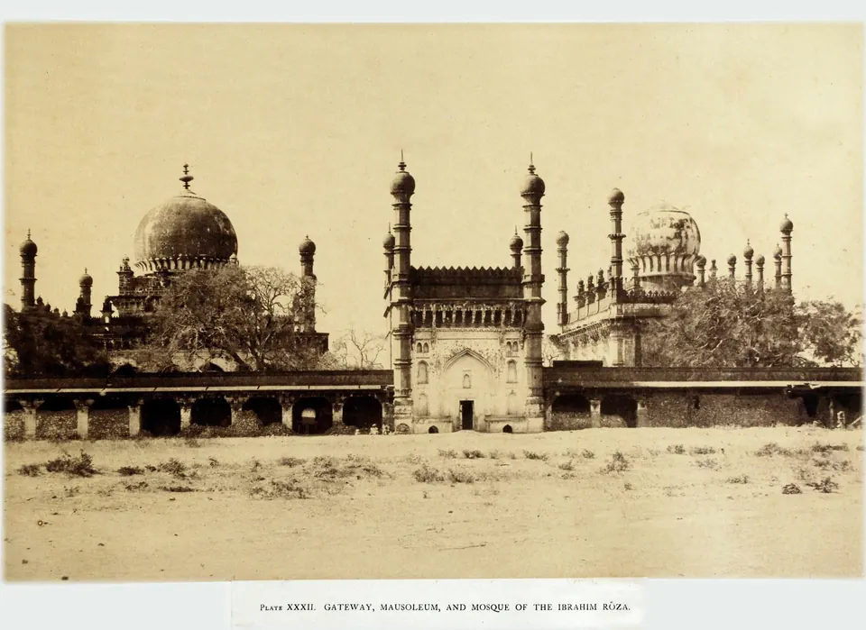 Sepia photograph of a walled enclosure seen across open ground, a tall gateway with minarets at the centre flanked by a domed tomb on the left and a mosque on the right.