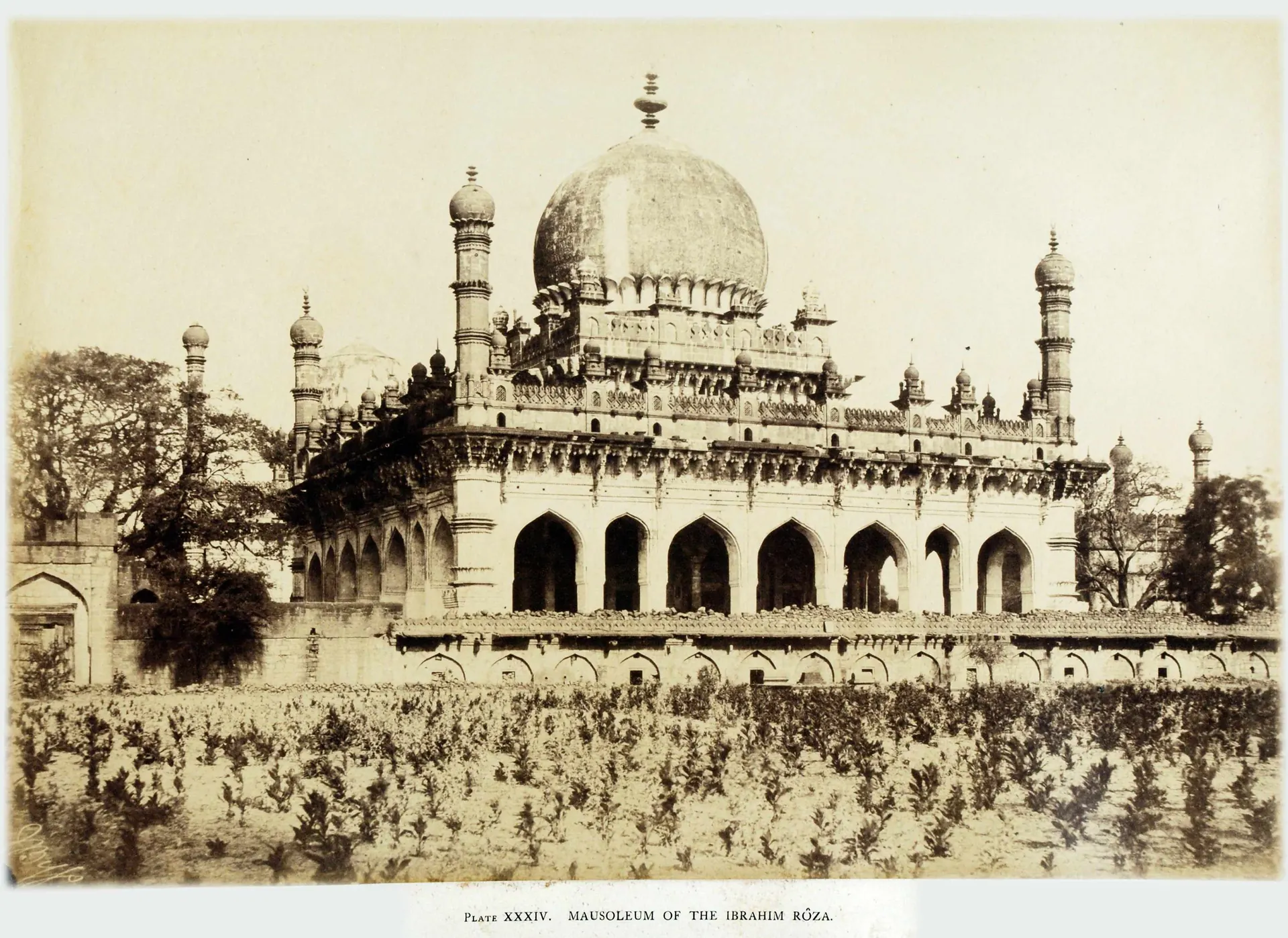 Sepia photograph of an ornate domed tomb on a high plinth, with arcaded verandahs, slender minarets and a bulbous dome on a petalled drum, behind a field of low shrubs.