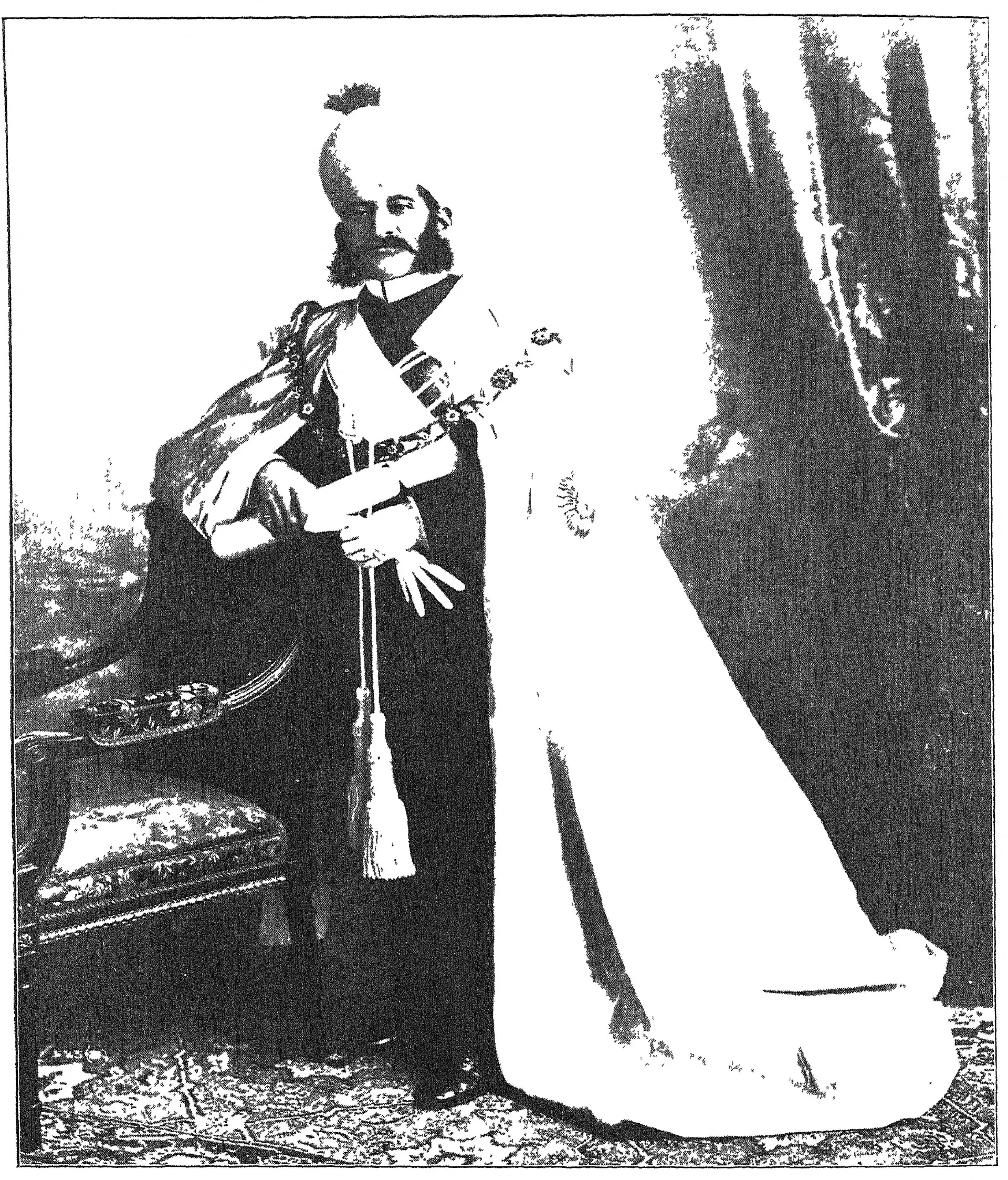 Photograph of Mahbub Ali Khan standing full length in a long white coat and a tall turban, with a sword.