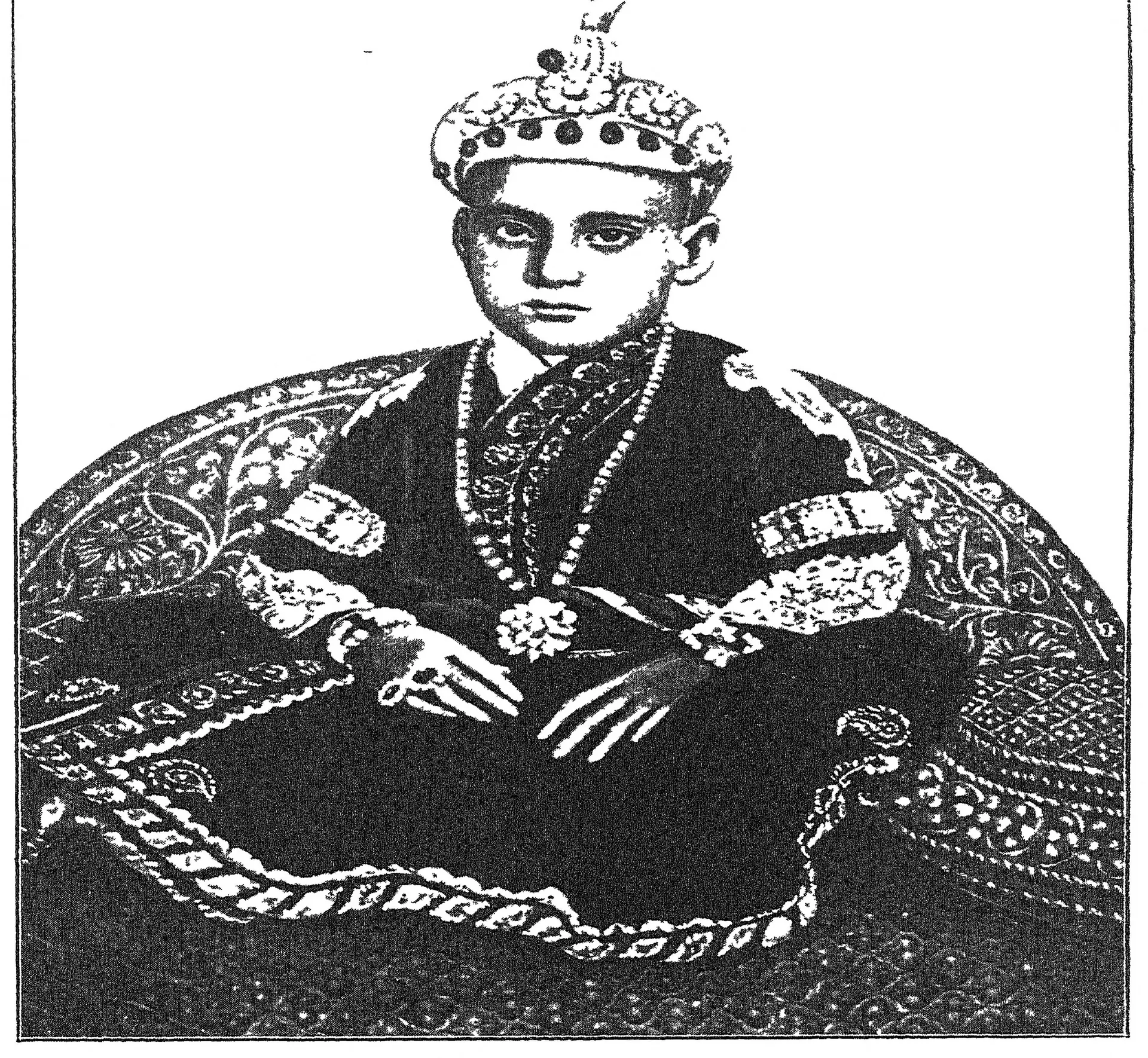 Photograph of the boy Nizam Mahbub Ali Khan seated cross-legged on a carpet with cushions, wearing a jewelled cap and robe.