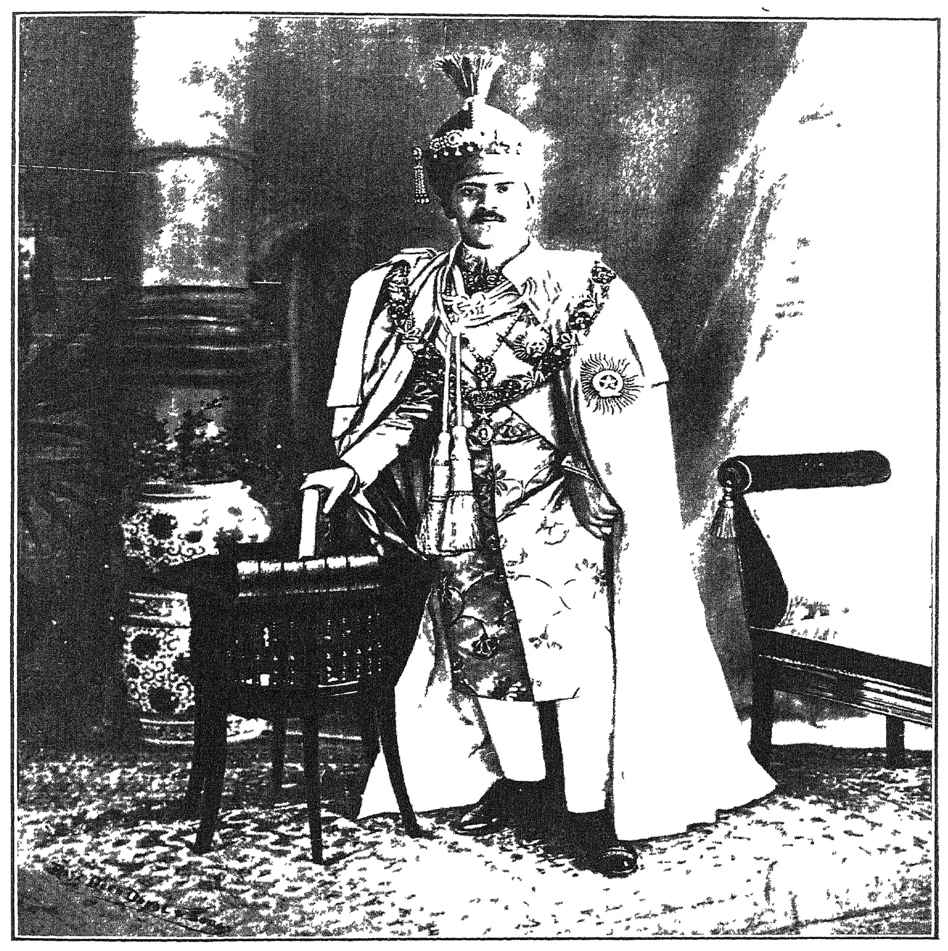 Photograph of Mir Osman Ali Khan standing in a richly embroidered coat and turban, beside a chair and a vase on a pedestal.