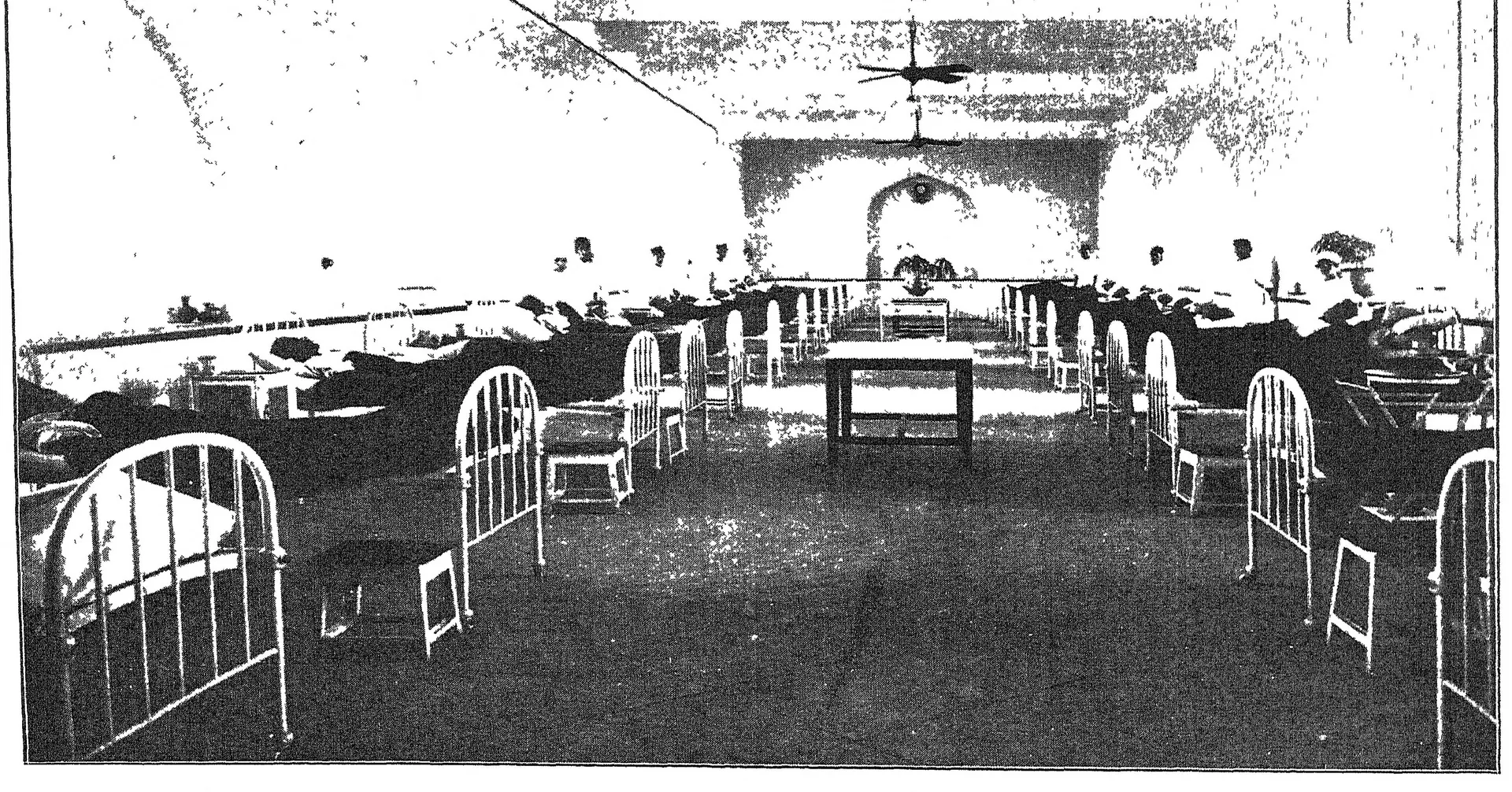 Photograph of a long hospital ward with rows of iron beds under a high arched ceiling with fans.