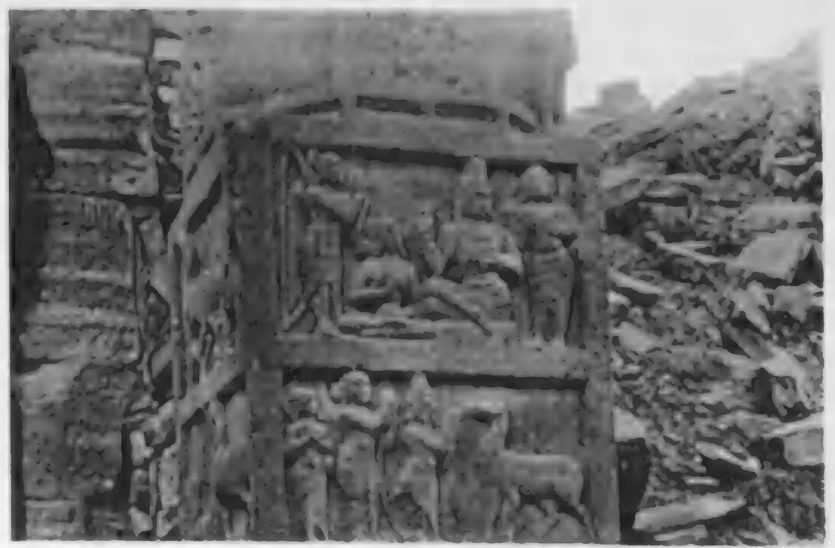 Old photograph of carved panels on a stone post: seated and standing figures in framed scenes.