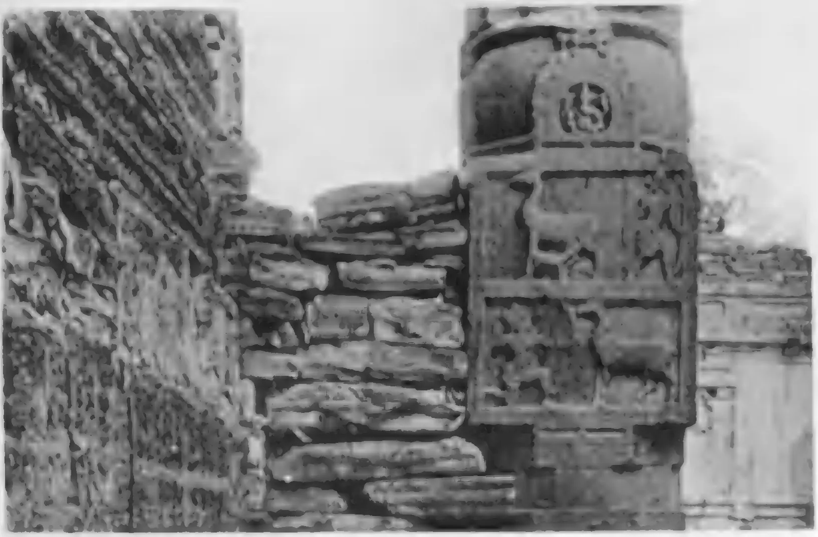 Old photograph of a carved stone gateway post with panels of small figures, standing among broken masonry.