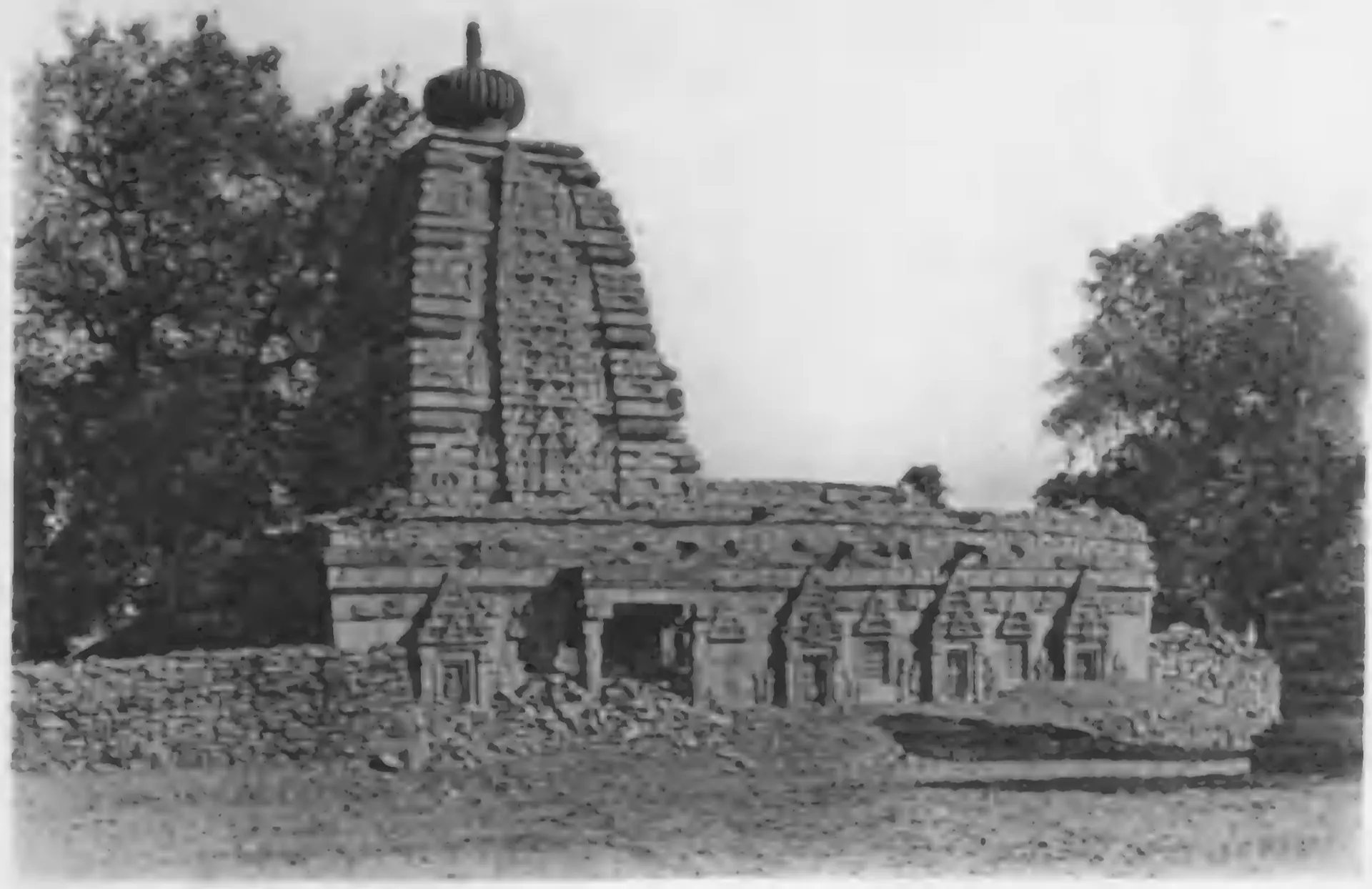 Old photograph of a stone temple with a tall curved tower crowned by a round ribbed stone, its long flat-roofed hall decorated with small niches, behind a rubble wall, with trees on both sides.
