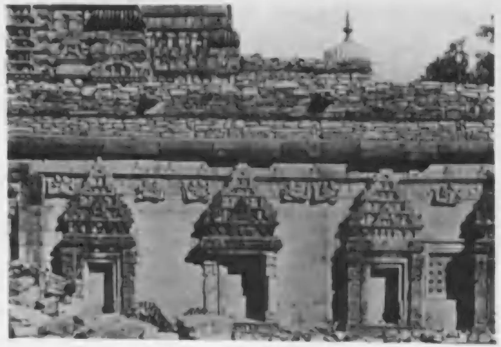 Old photograph of the carved outer wall of a temple, niches with small pediments above a moulded base, the tower top visible behind.
