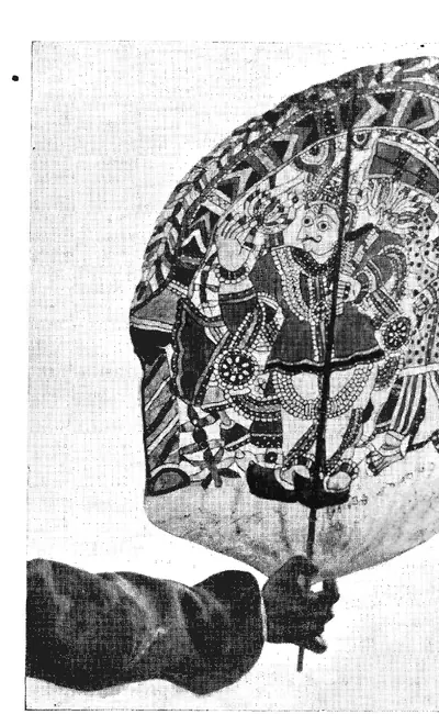 Black-and-white photograph of a hand holding up a large rounded leather puppet panel by a stick, painted with several crowned figures.