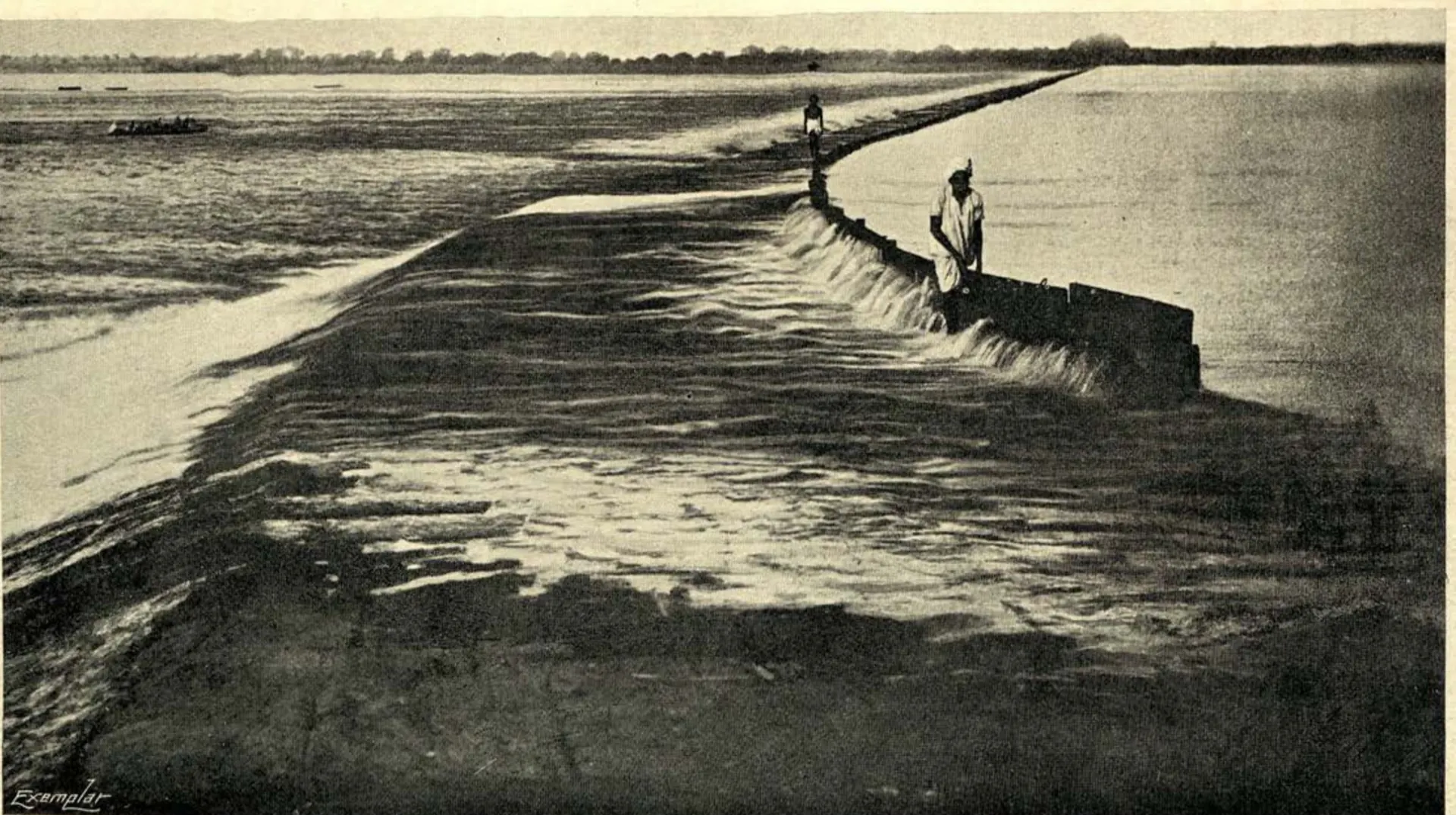 Old photograph of water pouring over a long low masonry dam across a wide river, with two men standing on the dam.