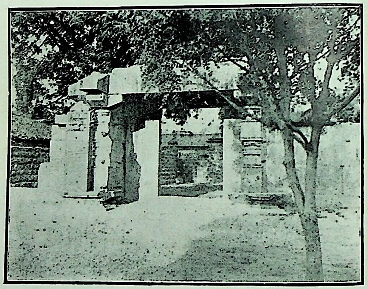 Old photograph of a stone gateway with a flat lintel among trees, a tall plain stone pillar standing to its left, and a dusty open ground in front.
