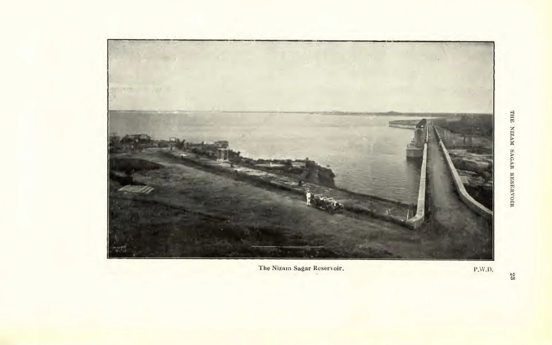 Photograph of the Nizam Sagar dam with the reservoir water on one side and buildings on the bank. Printed sideways.