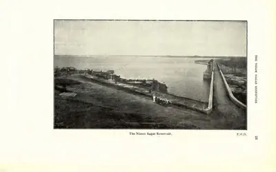 Photograph of the Nizam Sagar dam with the reservoir water on one side and buildings on the bank. Printed sideways.