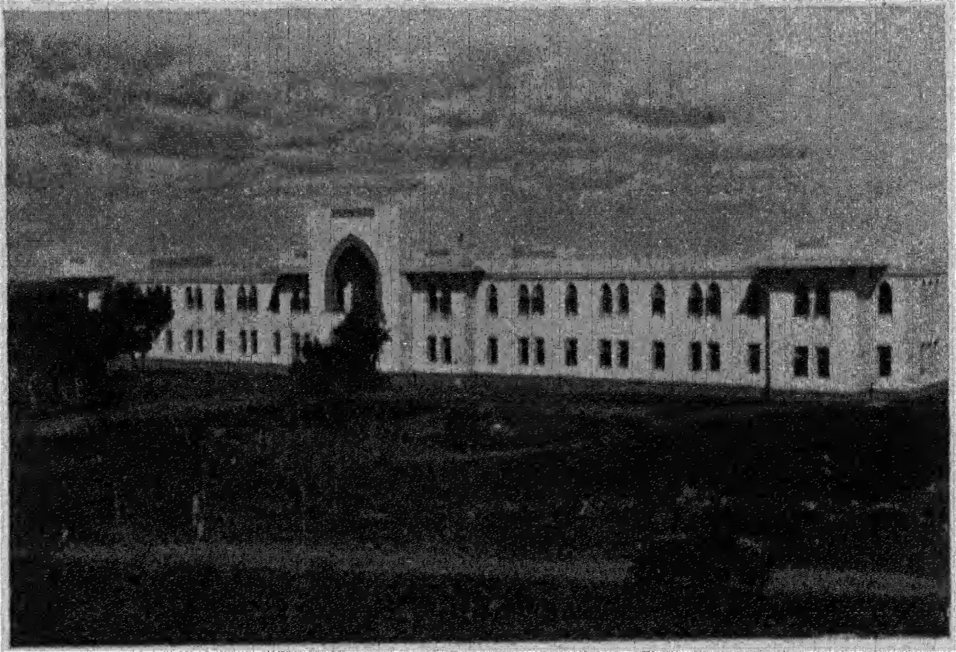 Photograph of a long two-storeyed hostel building with a tall pointed-arch entrance in the middle, seen across open ground.