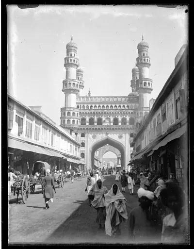 1887 glass-plate photograph of the Charminar at the end of a market street, with pedestrians in the foreground.