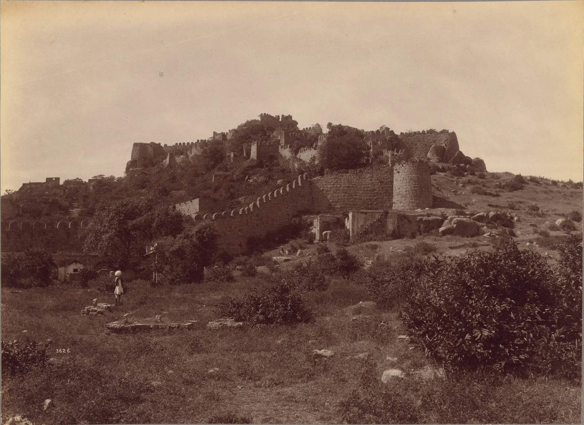1887 sepia albumen photograph of Golconda Fort's ruined walls across a scrubby hillside, with two men standing near the fort.