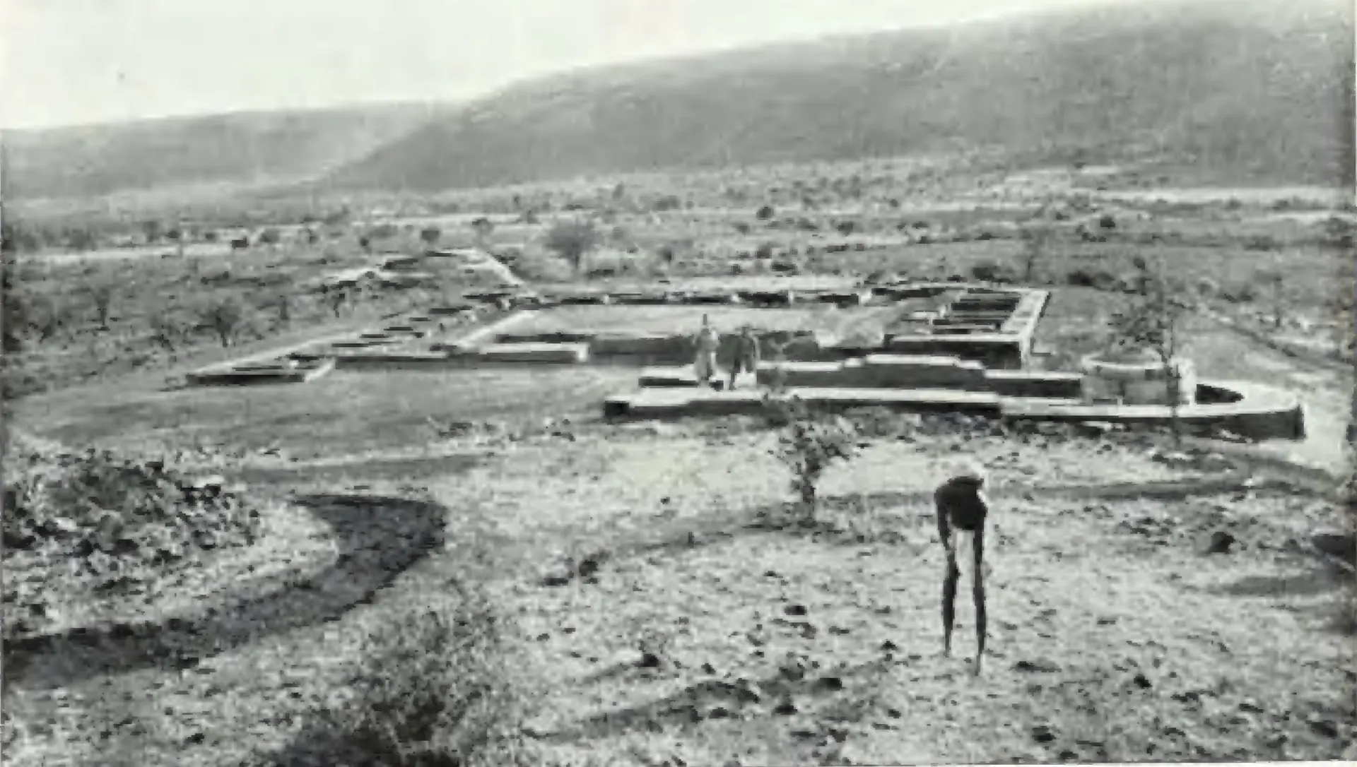Excavated brick foundations of a monastery and an apsidal temple on a wide valley floor, with hills behind and a man standing in the trench.