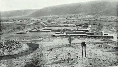 Excavated brick foundations of a monastery and an apsidal temple on a wide valley floor, with hills behind and a man standing in the trench.