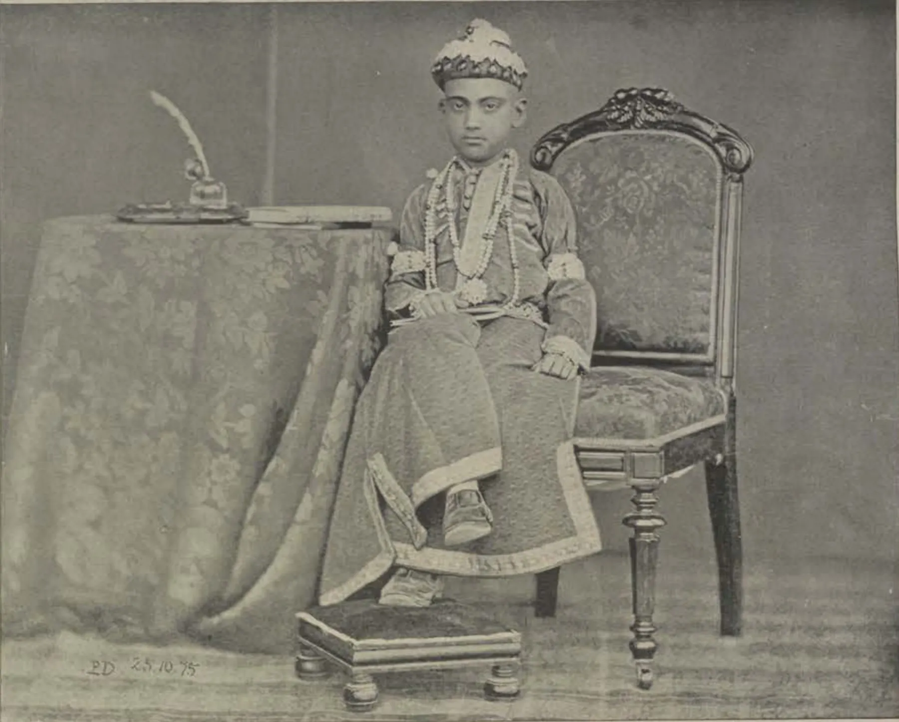 The young Nizam seated in an armchair beside a draped table, in an embroidered robe and a jewelled cap.