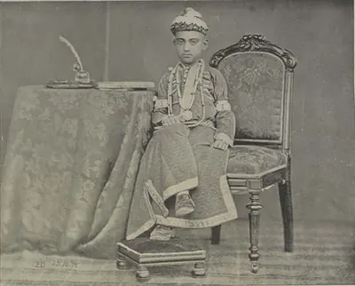 The young Nizam seated in an armchair beside a draped table, in an embroidered robe and a jewelled cap.