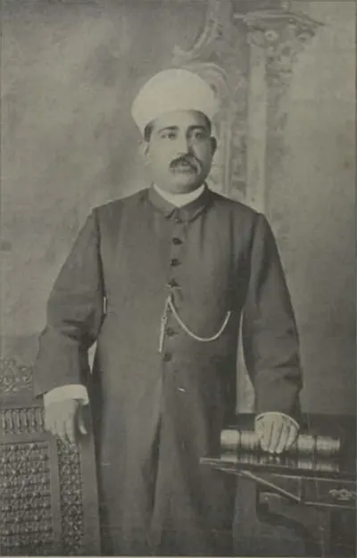 Syed Ali Bilgrami standing in a long buttoned coat and turban, one hand resting on books.
