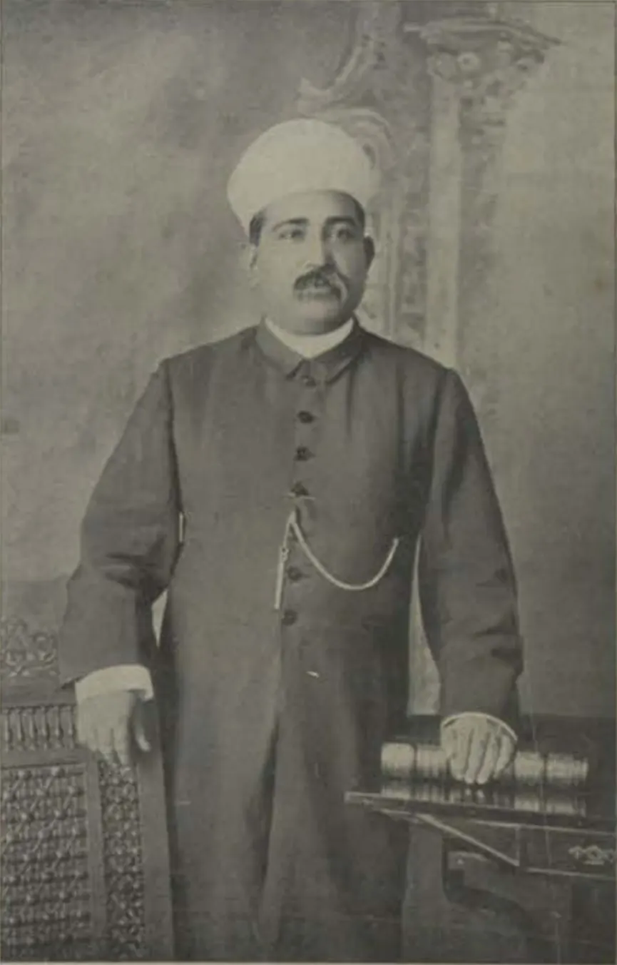 Syed Ali Bilgrami standing in a long buttoned coat and turban, one hand resting on books.