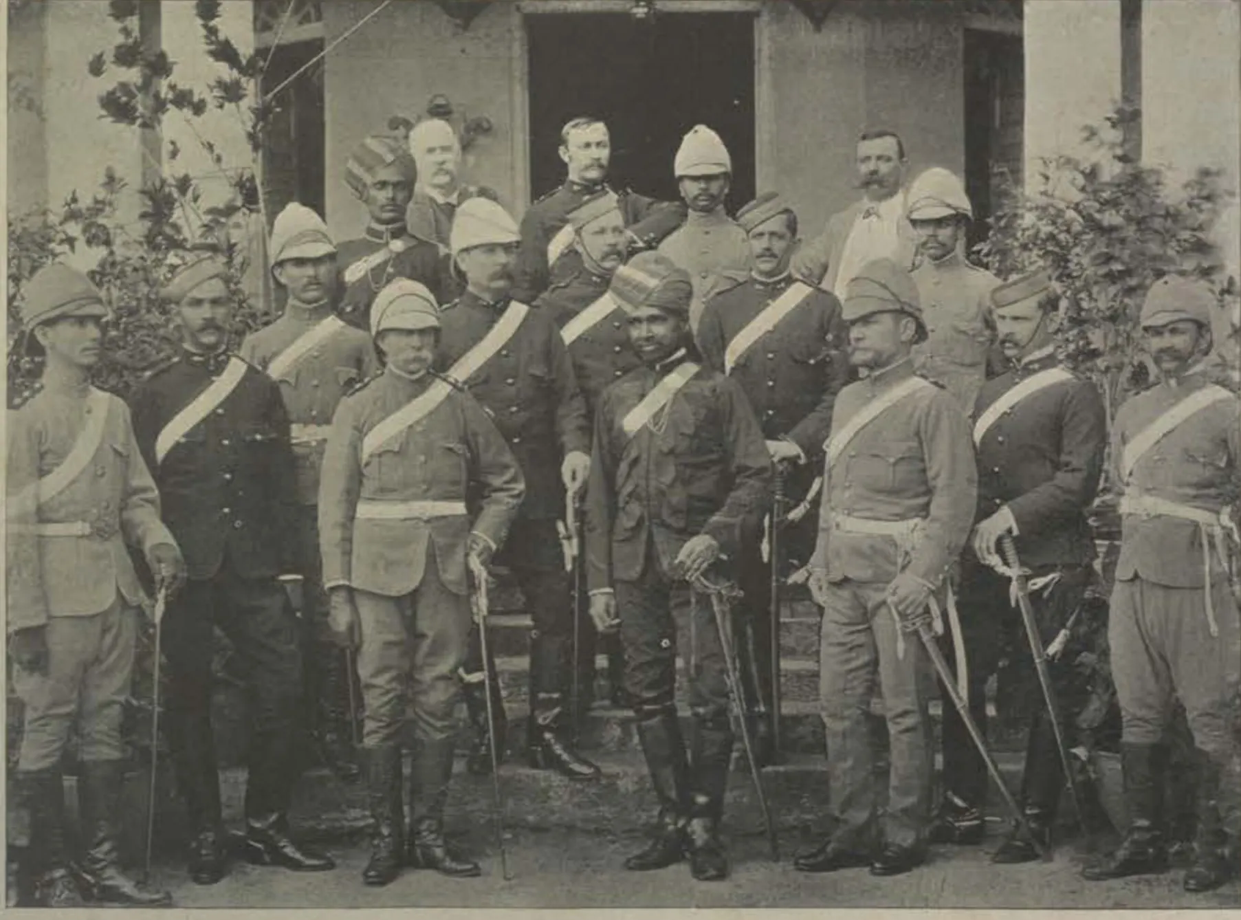Group of European and Indian officers in uniform, with helmets, turbans and swords, standing in front of a building.