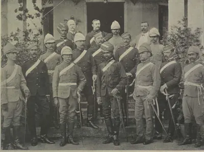 Group of European and Indian officers in uniform, with helmets, turbans and swords, standing in front of a building.