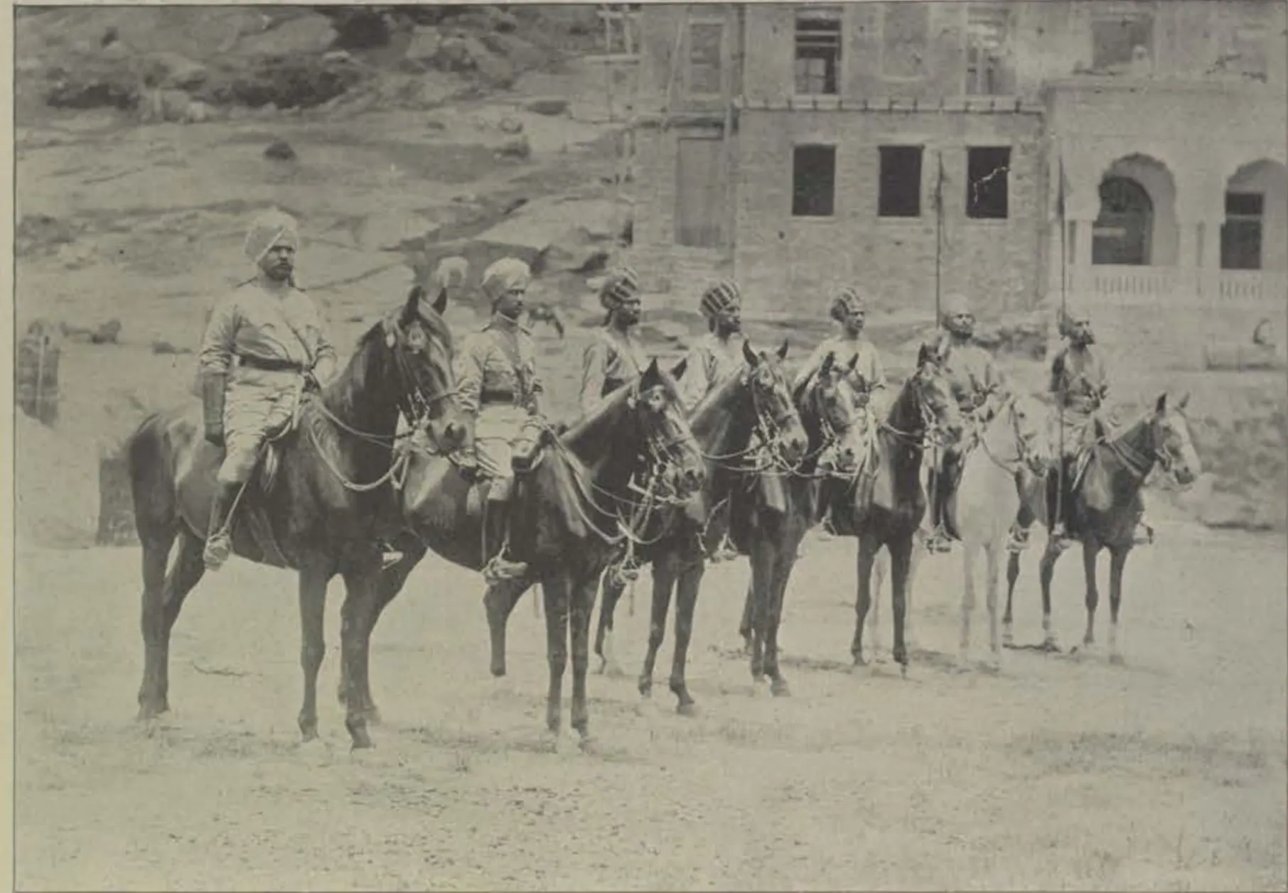 A line of mounted lancers in uniform, with a building behind.