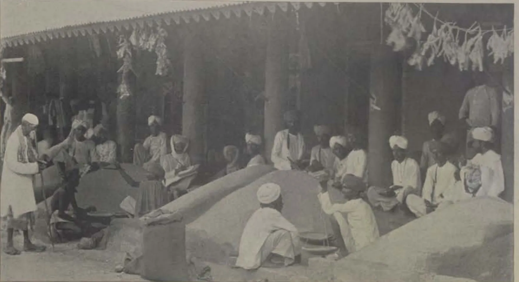 Traders seated beside heaps of grain under a roofed market.