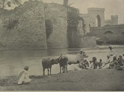A river bank with men and cattle, and ruined walls and a gateway beyond.