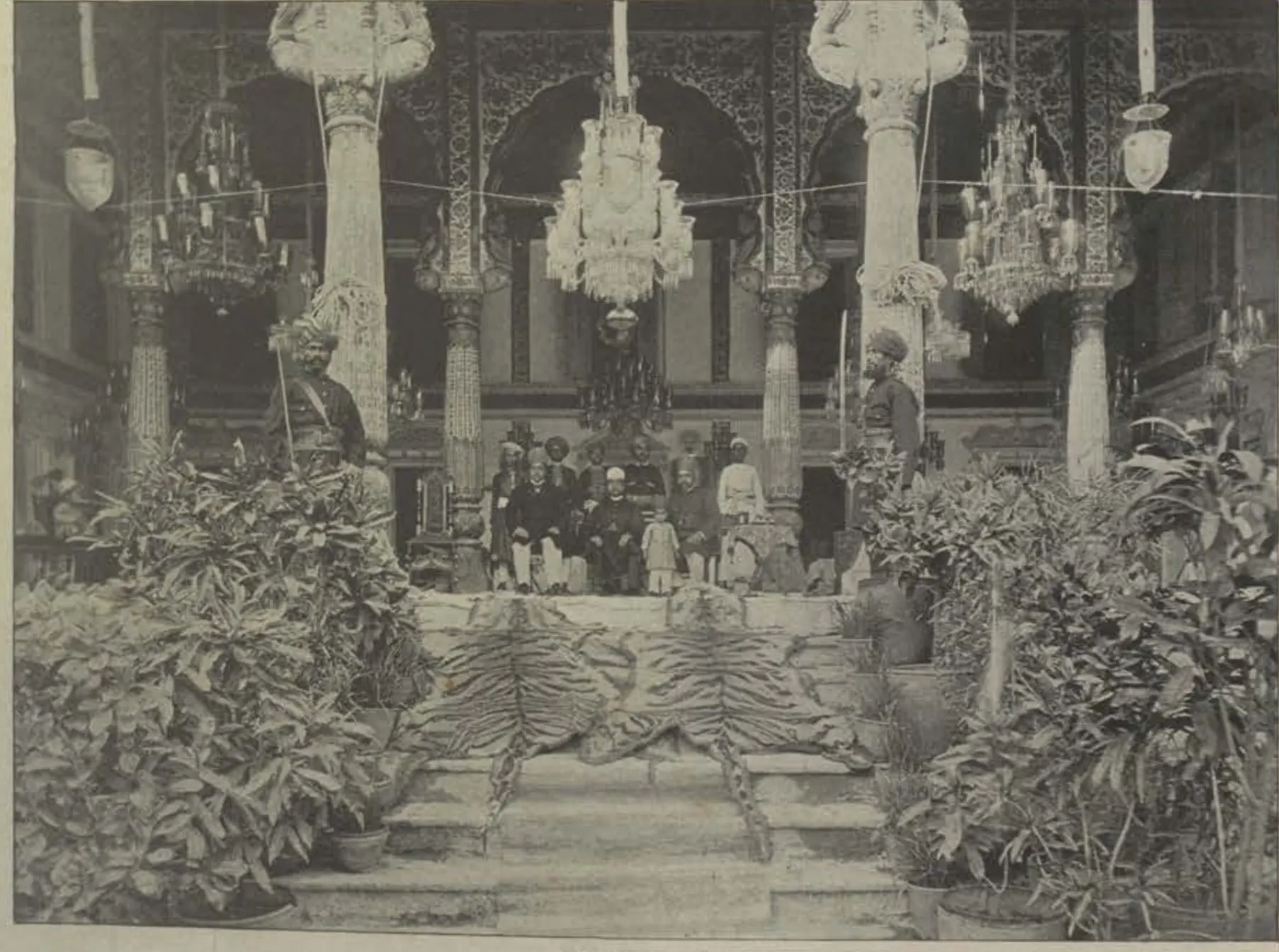 A hall of the Malwala palace with chandeliers, carved columns and potted plants.