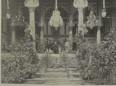 A hall of the Malwala palace with chandeliers, carved columns and potted plants.