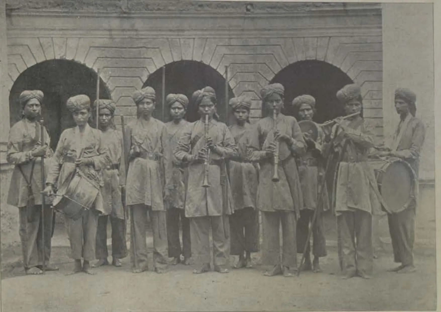 A line of women guards in uniform in front of an arcade, some holding a drum and musical instruments.