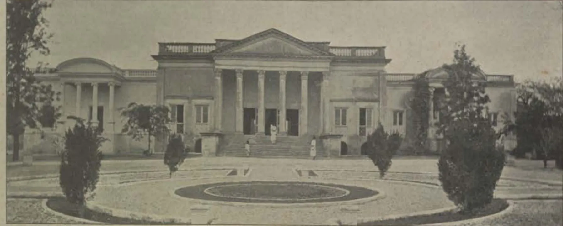 A classical building with a portico behind a circular lawn.