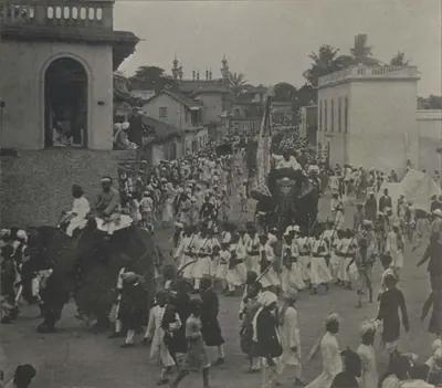 A standard and an elephant in a crowded street procession, with men marching.