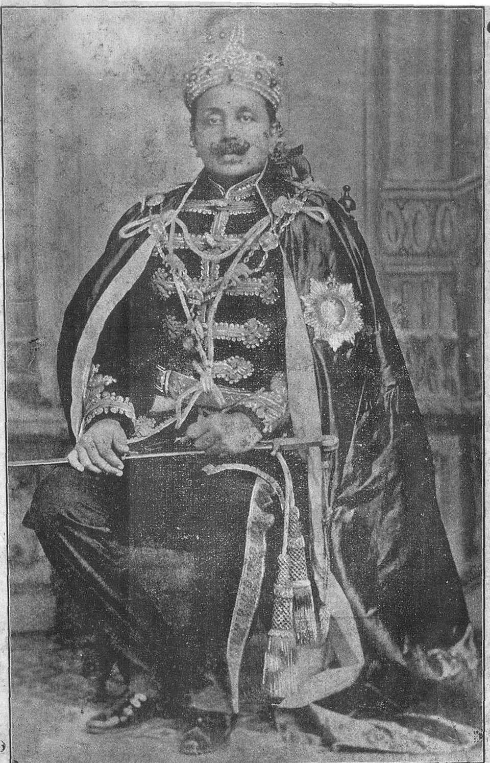 Old halftone photograph of a seated man with a moustache in a jewelled crown, an embroidered coat with braided frogging, a long velvet robe and a star, a sword across his lap.
