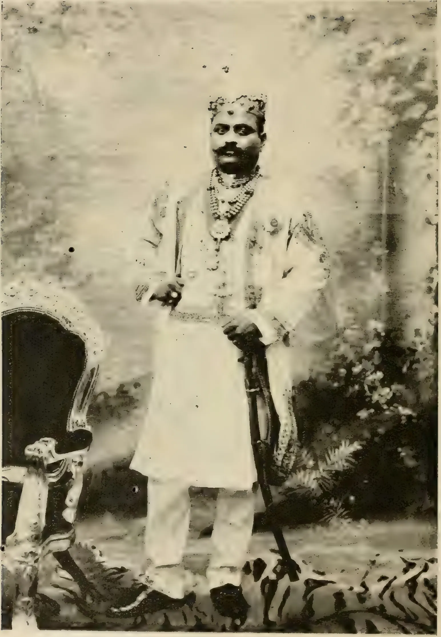 Old halftone photograph of a young man with a moustache, in a jewelled cap, pearl necklaces and a long white embroidered coat, standing with a sword beside a carved chair on a tiger skin before a painted garden backdrop.