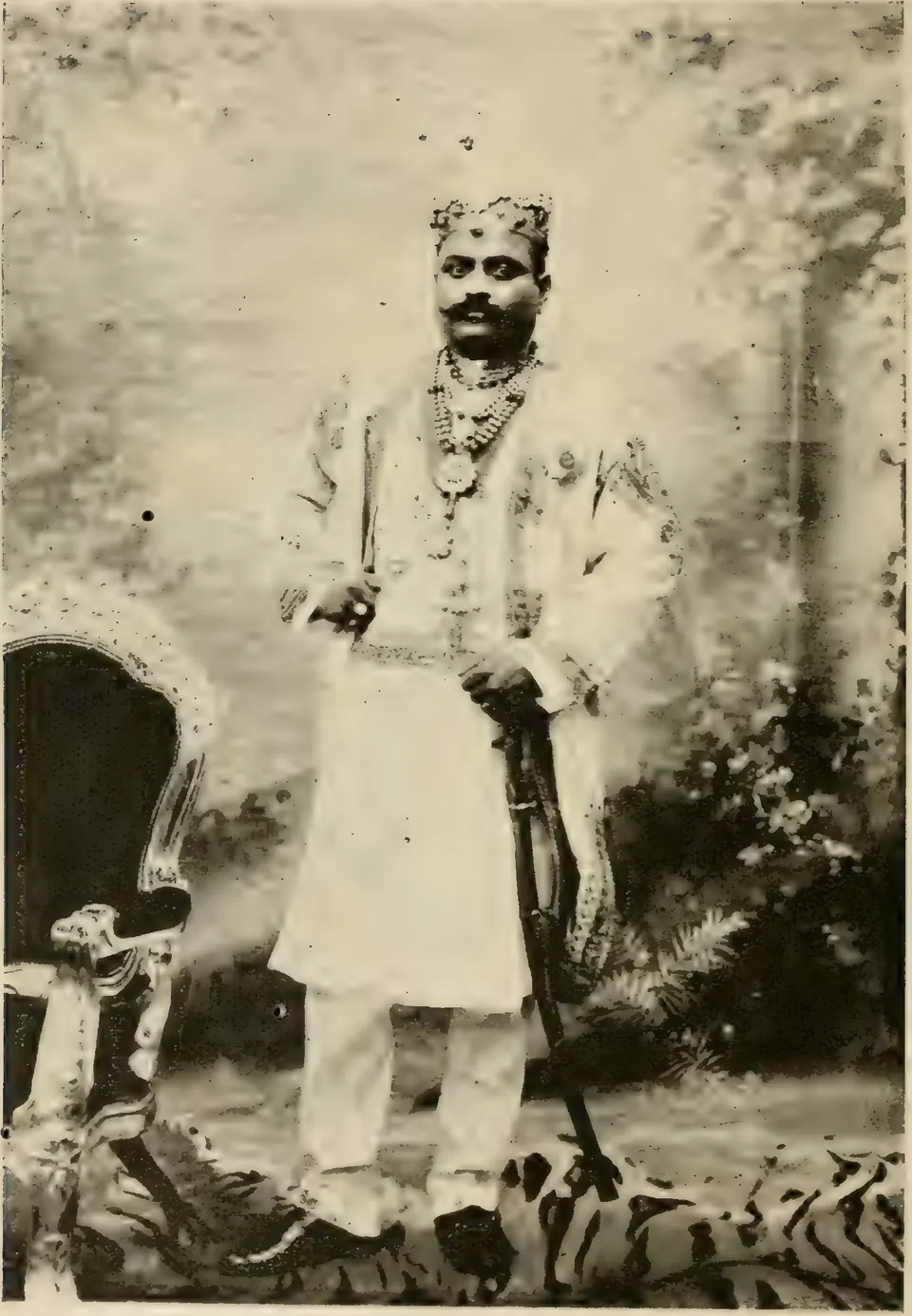 Old halftone photograph of a young man with a moustache, in a jewelled cap, pearl necklaces and a long white embroidered coat, standing with a sword beside a carved chair on a tiger skin before a painted garden backdrop.