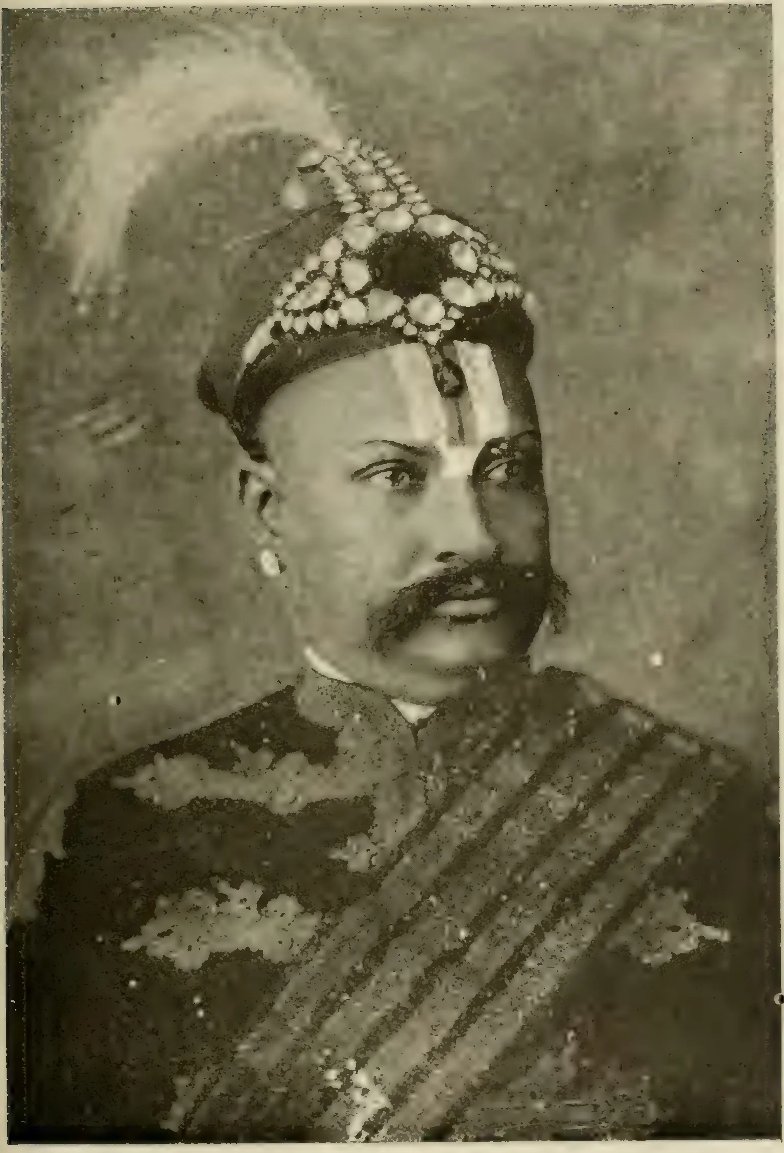 Old halftone head-and-shoulders photograph of a man with a large moustache and a caste mark, wearing a jewelled turban with a plume and a dark coat with a striped sash.