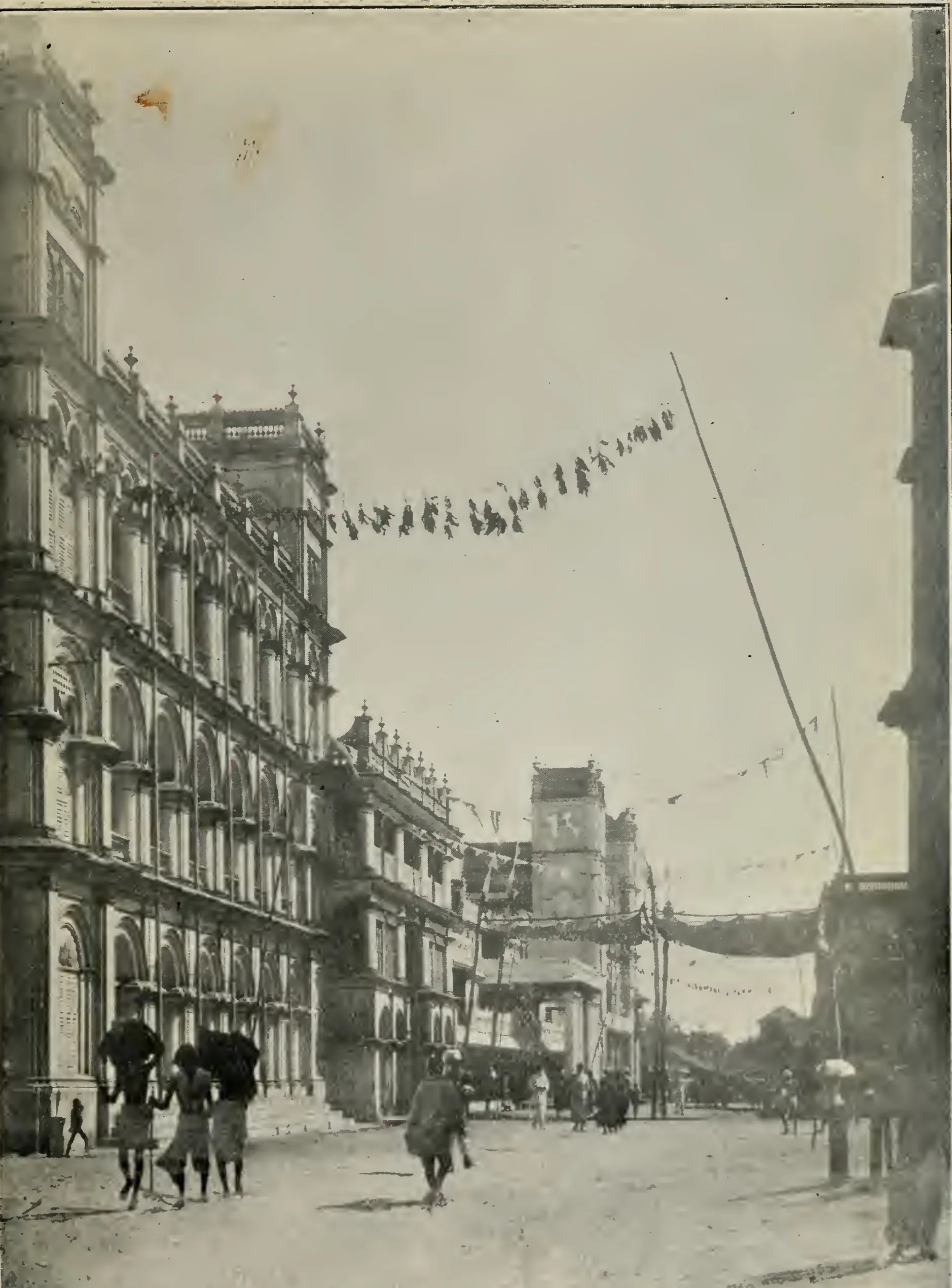Old photograph of a street with a long three-storeyed palace façade of arched windows and balconies on the left, flags strung across the road on a pole, and people walking with umbrellas.
