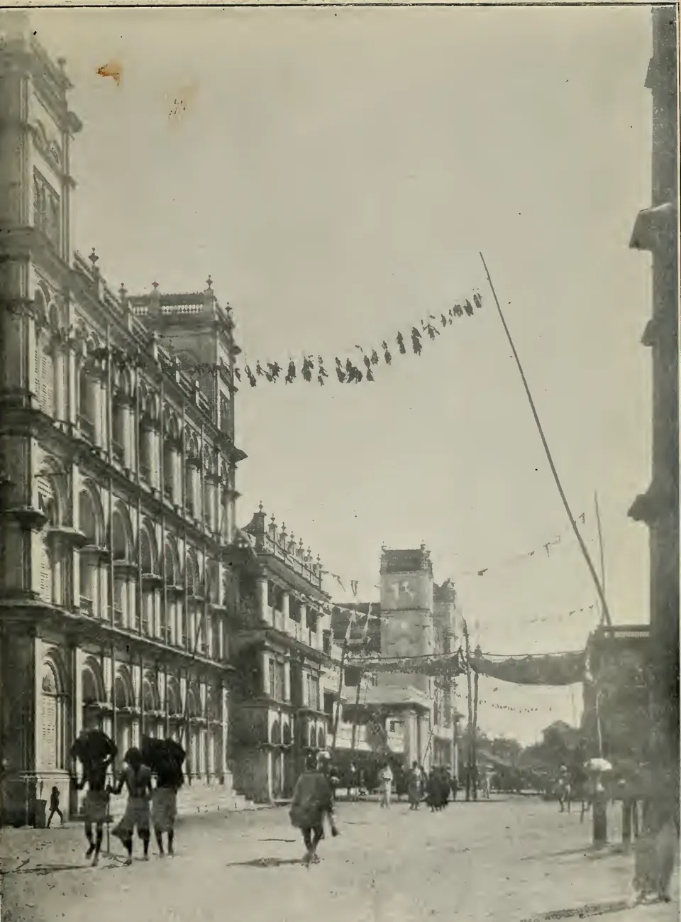 Old photograph of a street with a long three-storeyed palace façade of arched windows and balconies on the left, flags strung across the road on a pole, and people walking with umbrellas.