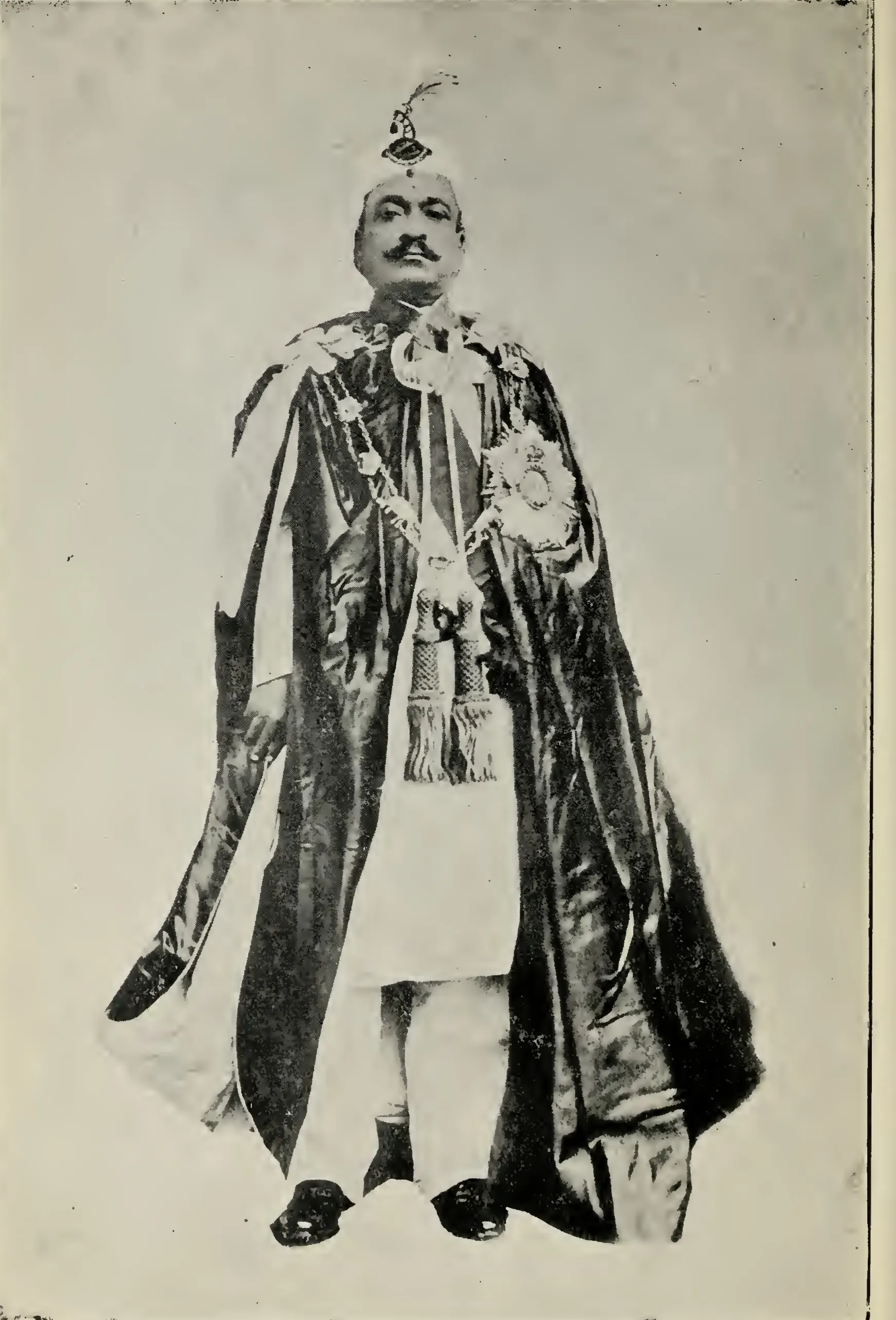 Old halftone photograph of a man with a moustache standing in a plumed turban and a long dark robe of state with a broad sash, collar and star, over white trousers.