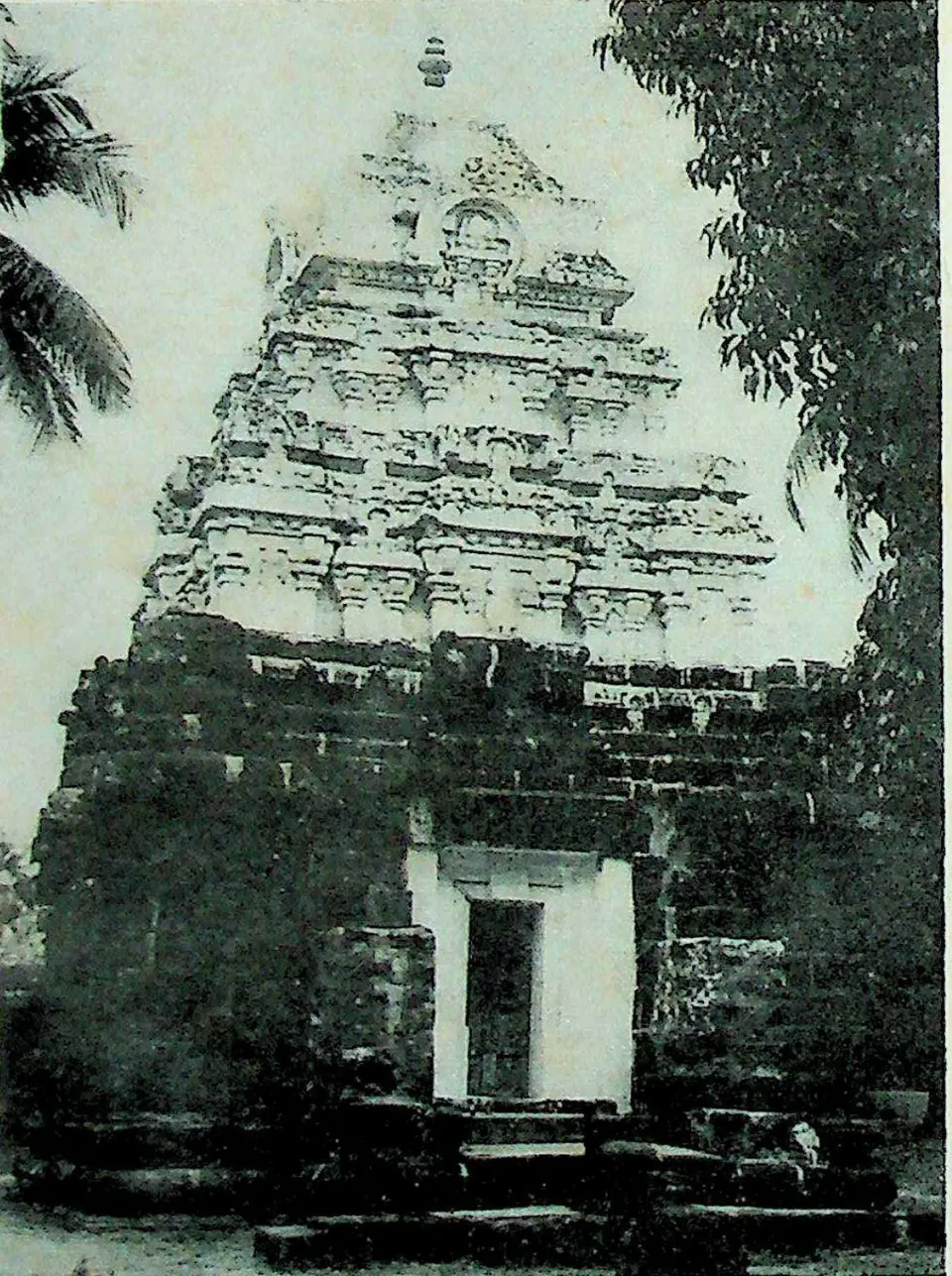 A small square stone shrine with a dark, weathered wall of large blocks and a plain doorway, topped by a stepped pyramidal tower of carved storeys; palm fronds at the left and a tree at the right.
