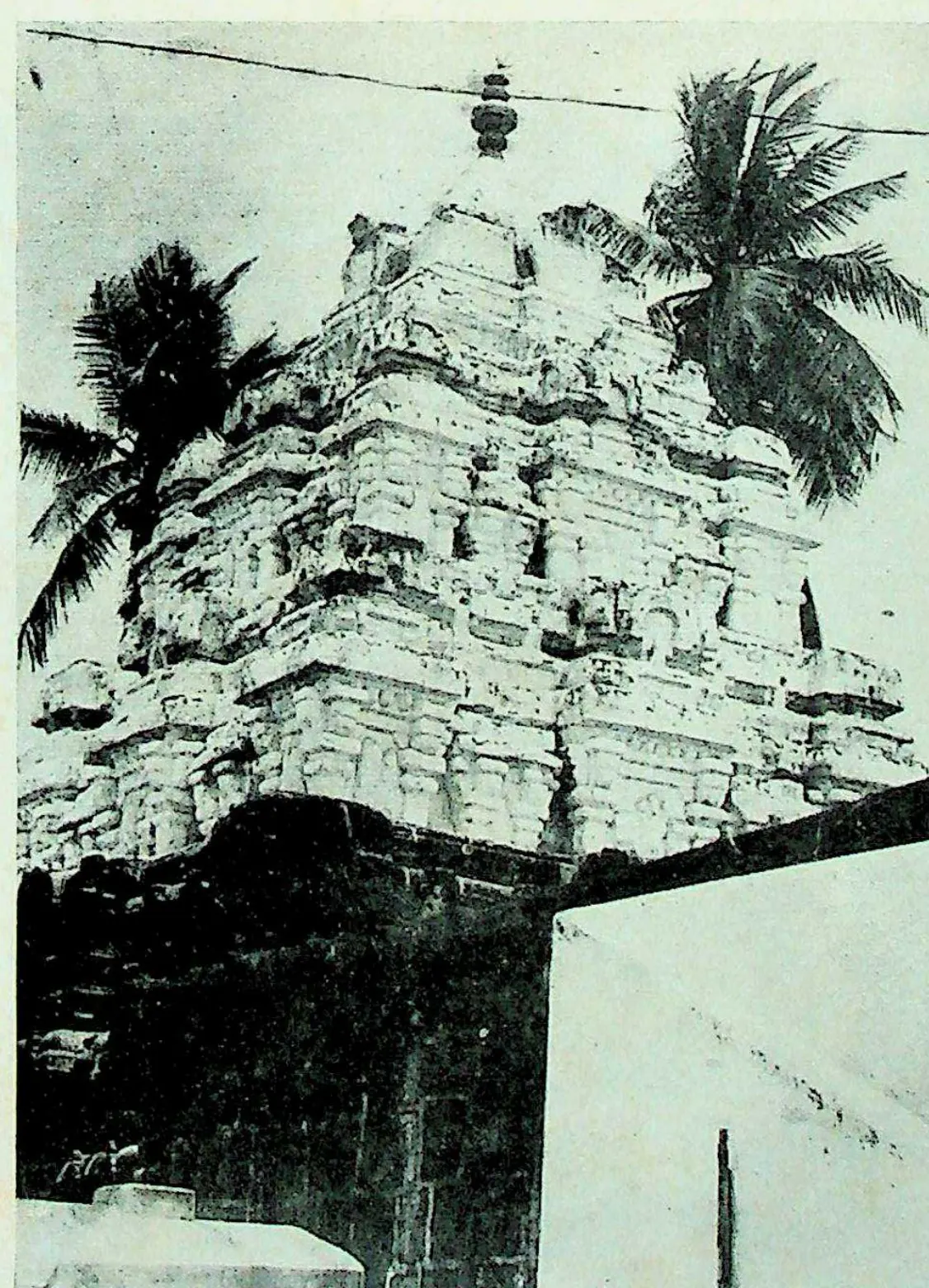 The tiered stone tower of a shrine, its storeys crowded with carved niches and miniature roofs, rising behind a dark flat-roofed porch, with coconut palms and an overhead wire behind.