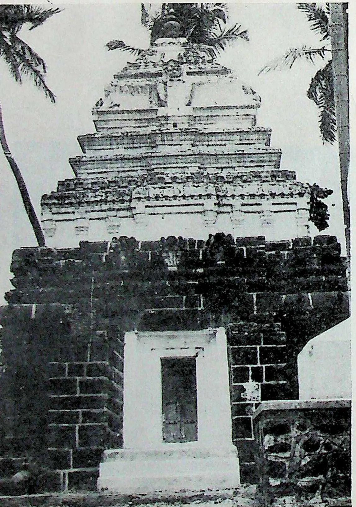 A square shrine of dark masonry with a whitewashed doorway frame, crowned by a pale stepped tower of four receding storeys; palms on both sides.