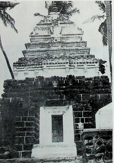 A square shrine of dark masonry with a whitewashed doorway frame, crowned by a pale stepped tower of four receding storeys; palms on both sides.