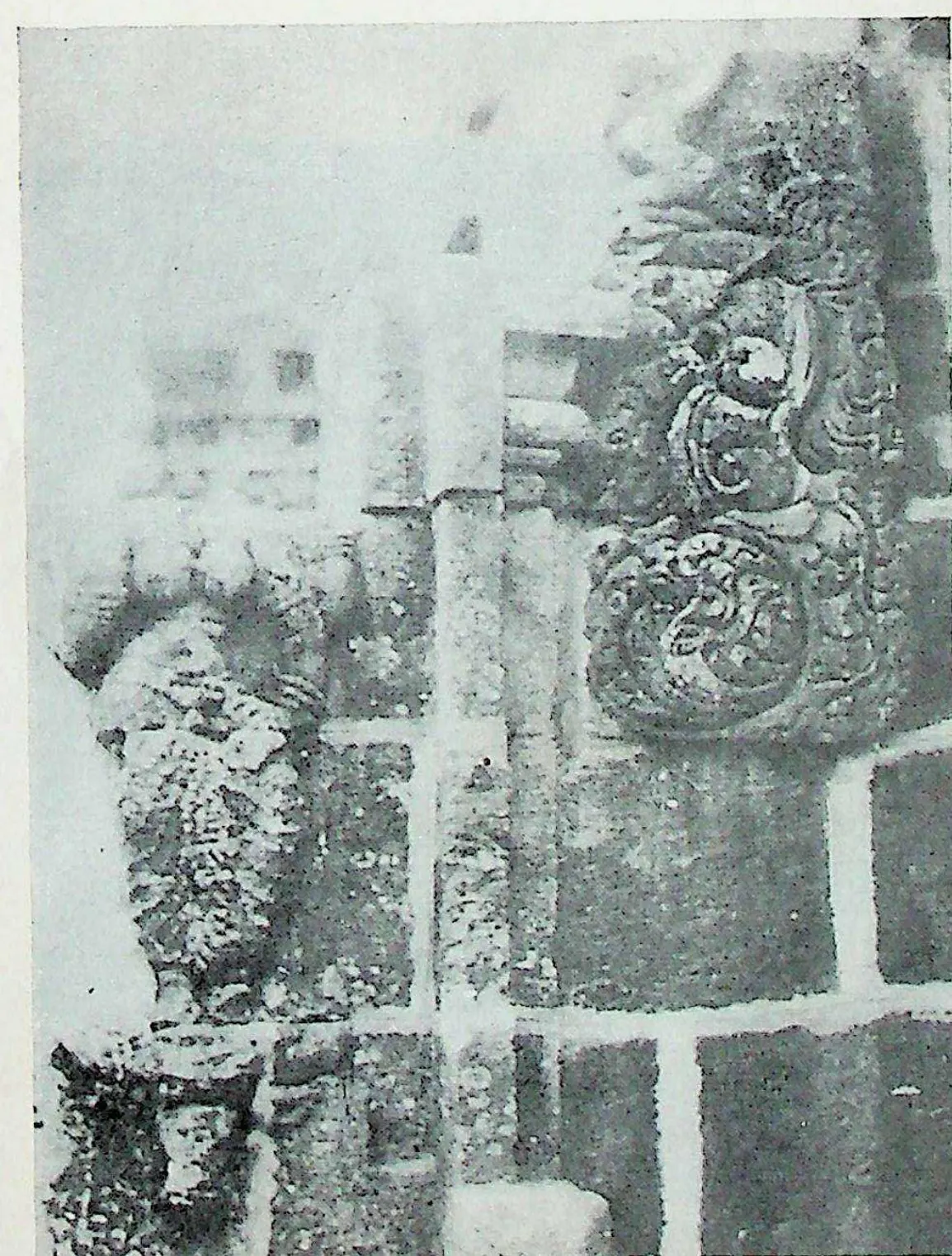 Close view of a temple wall projection: stacked stone blocks with a carved scroll-work panel and a pilaster with a pot-and-foliage capital; much of the surface is pale and worn.