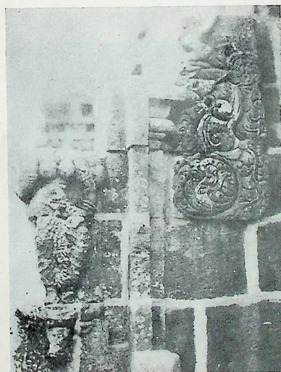 Close view of a temple wall projection: stacked stone blocks with a carved scroll-work panel and a pilaster with a pot-and-foliage capital; much of the surface is pale and worn.
