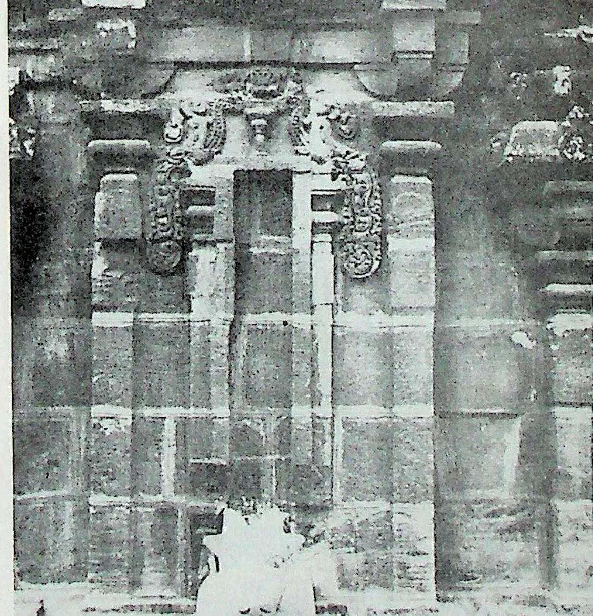 The side wall of a stone temple with a central niche framed by pilasters and curling makara-arch ornament, under a cornice of miniature roofs; fallen material at the foot.
